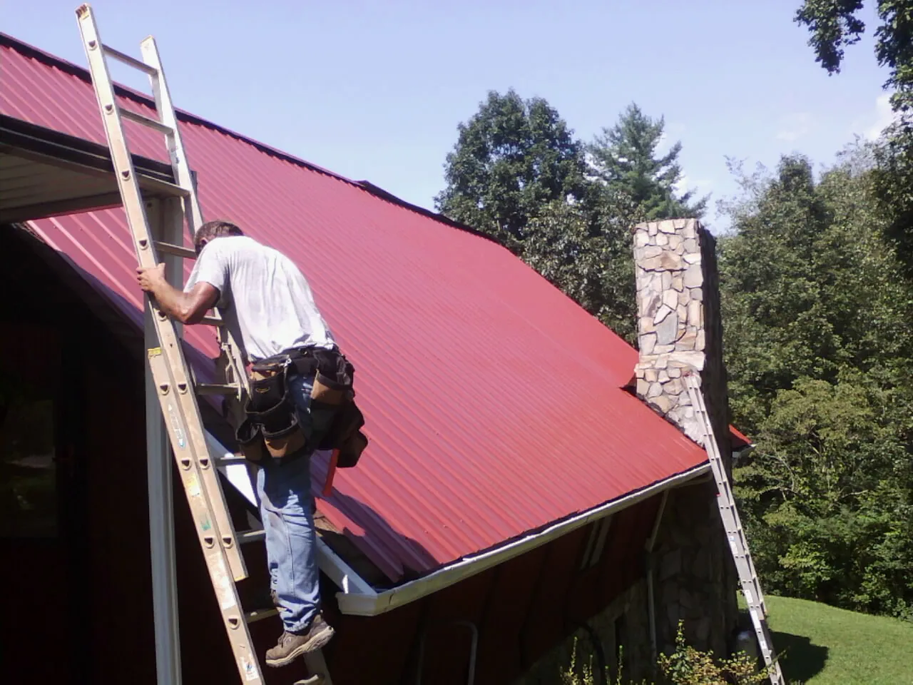 Roofer on a ladder beside a red metal roof and stone chimney — Soffit & Fascia Repair by Roofing Brothers in Summit Argo